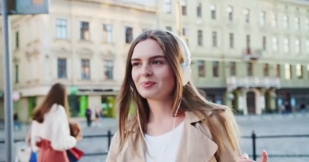 Close up portrait of joyful Caucasian young female on street listening to music in headphones. Beautiful happy girl dancing while walking in city and enjoying songs outdoor. Leisure concept - Metraje, vídeo
