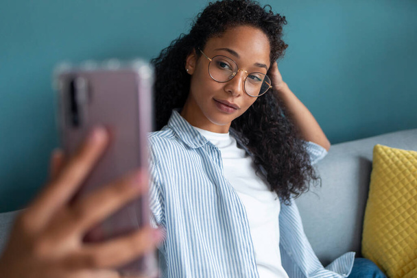 Shot van interessante jonge Afrikaanse Amerikaanse vrouw met behulp van haar mobiele telefoon om een selfie te nemen terwijl zitten op de bank thuis. - Foto, afbeelding