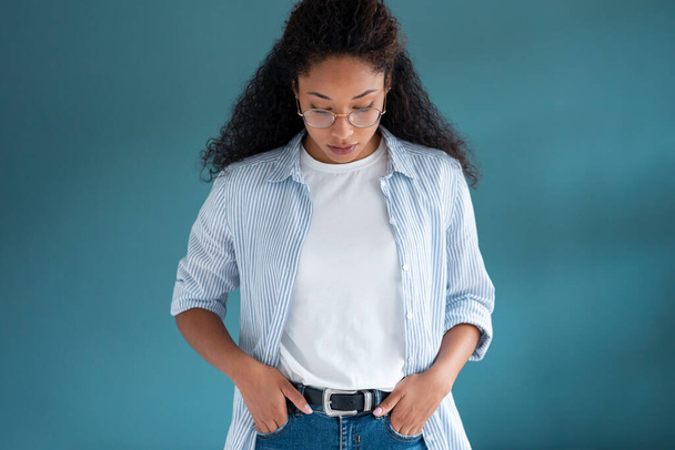 Portret van mooie jonge Afrikaanse Amerikaanse vrouw op zoek naar beneden met serieuze expressie terwijl poseren over blauwe achtergrond. - Foto, afbeelding