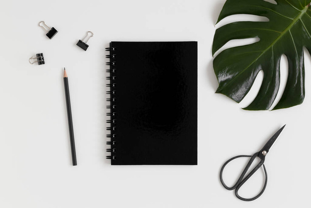 Draufsicht auf eine schwarze Notizbuch-Attrappe mit Arbeitsplatzzubehör und einem Monsterblatt auf einem weißen Tisch. - Foto, Bild