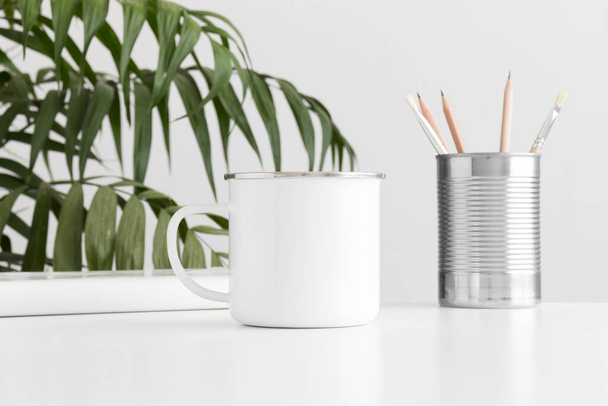 Taza de esmalte maqueta con accesorios de espacio de trabajo en una mesa blanca y una planta de palma
. - Foto, Imagen
