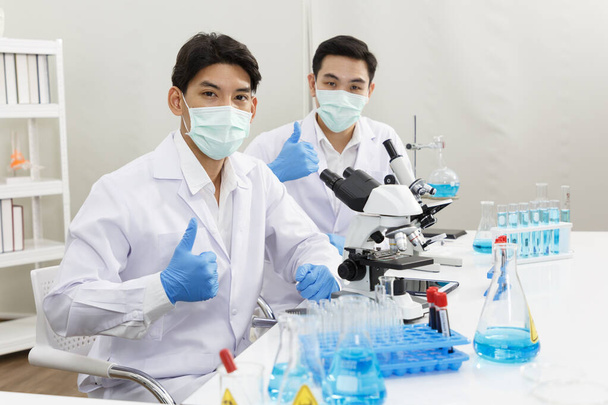 Zwei männliche Wissenschaftler tragen Gesichtsmaske bei der Arbeit im Labor, während sie Daumen hoch und bereit zur Arbeit und Präsentation zeigen. Blauton. SARS-CoV-2, Covid-19 THEME. - Foto, Bild