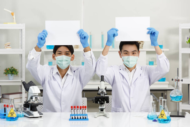 Двое ученых-мужчин носят маску для лица в лаборатории, держа в руках чистую бумагу. Скопируй пространство. SARS-CoV-2, Covid-19 THEME. - Фото, изображение