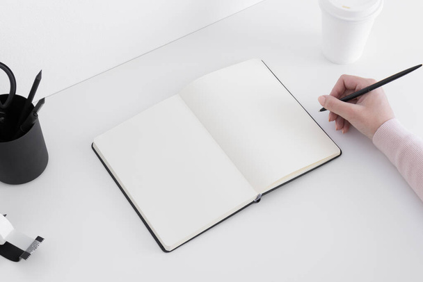 Open notebook mockup met een koffiebeker, werkruimte accessoires op een witte tafel en met vrouw die aantekeningen maakt. - Foto, afbeelding