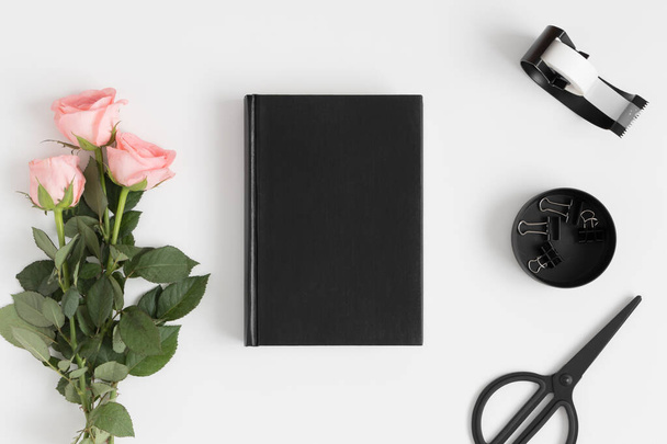 Top view of a black book mockup with a bouquet of pink roses and workspace accessories on a white table. - Foto, imagen
