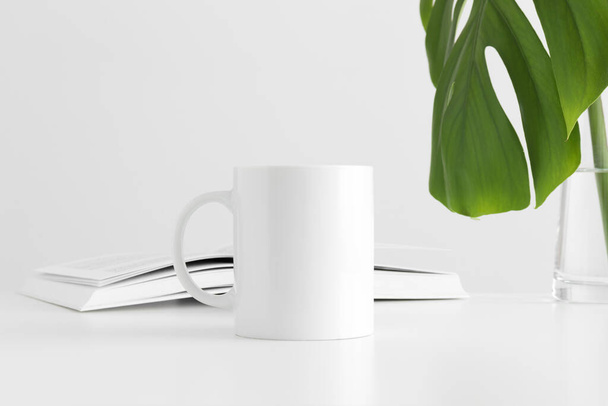 White mug mockup with a monstera leaf in a vase and a book on a white table. - Foto, imagen