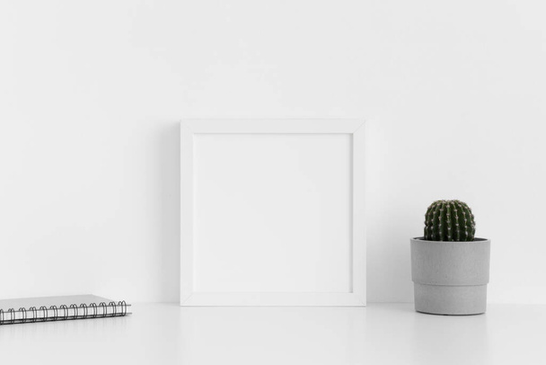 Weiße quadratische Rahmen-Attrappe mit einem Kaktus im Topf und Notizbuch auf einem weißen Tisch. - Foto, Bild