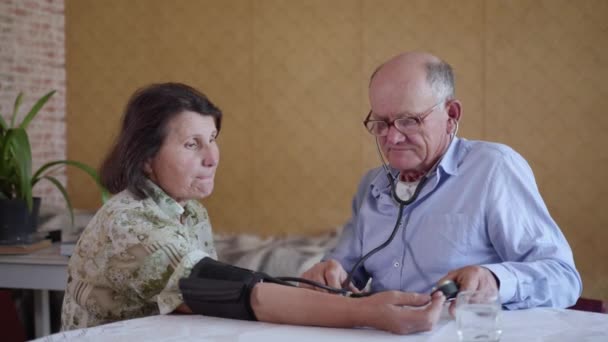 elderly caring man checks blood pressure of his old wife with hypertension using a blood pressure monitor while sitting at home at the table - Materiał filmowy, wideo