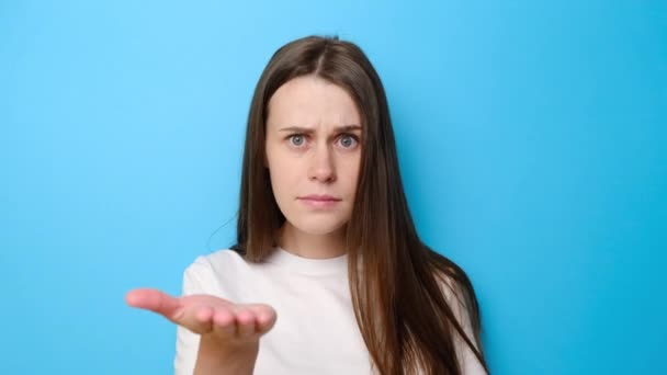 Annoyed young woman pointing at camera and showing stupid gesture, blaming some idiot, dressed in white t-shirt, isolated on blue studio background. Portrait of brunette girl in despair and shock - Filmagem, Vídeo