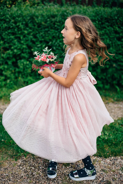 A little girl in a long dress is spinning and holding flowers in her hands, in the background is a garden. Holiday birthday. Style and fashion concept. Carefree childhood. - Photo, Image