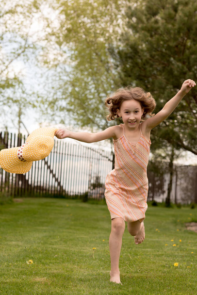 A little girl in a yellow dress with a hat runs in the summer in the country. The child runs a half-horse. Short shiny curly hair. The concept of children's power and fashion. Carefree childhood. - Photo, image
