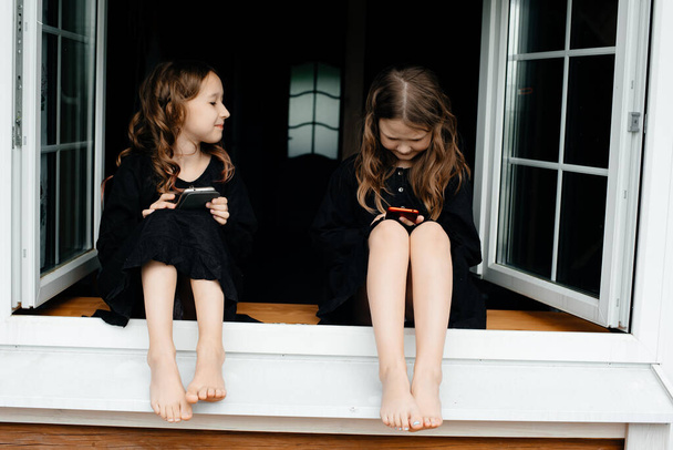 Little girls in black dresses are sitting at the window and talking on the phone, laughing and smiling. Long shiny dark hair. Summer holidays Carefree childhood.  - Φωτογραφία, εικόνα