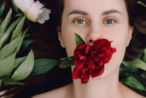 Top view close up beauty portrait of beautiful attractive young woman with red peony covering mouth, looking at camera. Natural cosmetics made of flower petals, clean delicate skin of girl face - 写真・画像