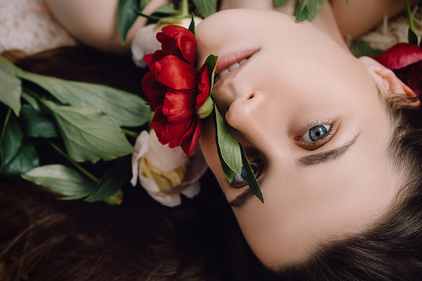 Side view of cute charming pretty young woman lying on hair with flower peony covering mouth. Beauty portrait of beautiful brunette girl looking at camera. Haircare, skin beauty health care concept - Valokuva, kuva