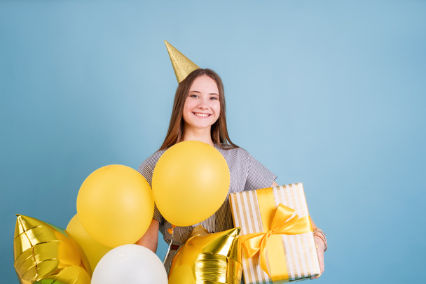Doğum günü partisi. Doğum günü şapkalı genç kadın elinde balonlar ve büyük hediye kutusuyla mavi arka planda doğum günü partisini fotokopi aletiyle kutluyor. - Fotoğraf, Görsel