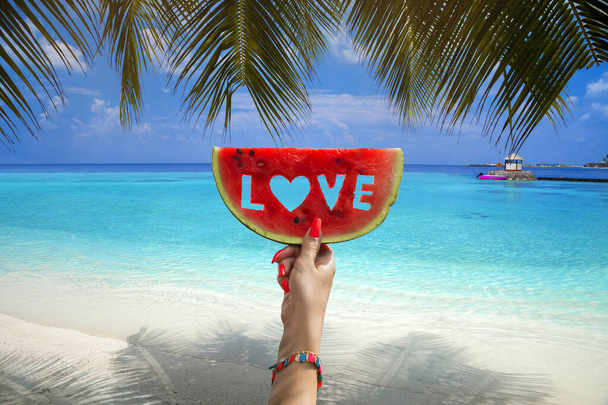 Watermeloen snijdt met tekstliefde, vrouwenhanden houden het tegen de blauwe lucht en de Indische oceaan. Zomer concept.Tropisch eiland strand. Malediven eiland. - Foto, afbeelding