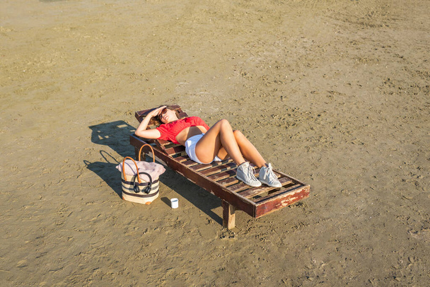 Beautiful girl in a shorts and blouse is relaxing on wooden lounge chair on a sandy beach by the sea. Top view. Sunset. - Φωτογραφία, εικόνα