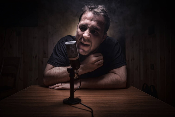 Caucasian man news broadcaster sat at a desk with microphone in dark room. Performer with microphone. Male musician singing into a microphone. Shot on a black background. Dark atmosphere - Photo, Image
