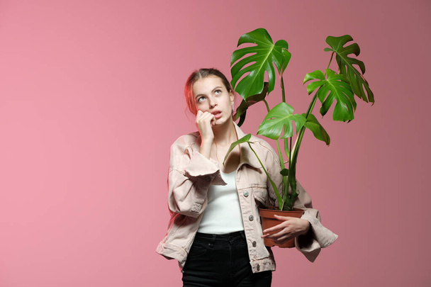 A girl in a pink jacket in black jeans holds in her hands a monstera flower in a pot posing on a pink background. - Valokuva, kuva