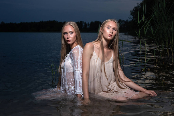 Две молодые сестры-близнецы позируют в светлых платьях в воде озера в летнюю ночь - Фото, изображение