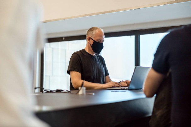 Человек в маске, работающий за ноутбуком в аэропорту в ожидании своего рейса - Фото, изображение