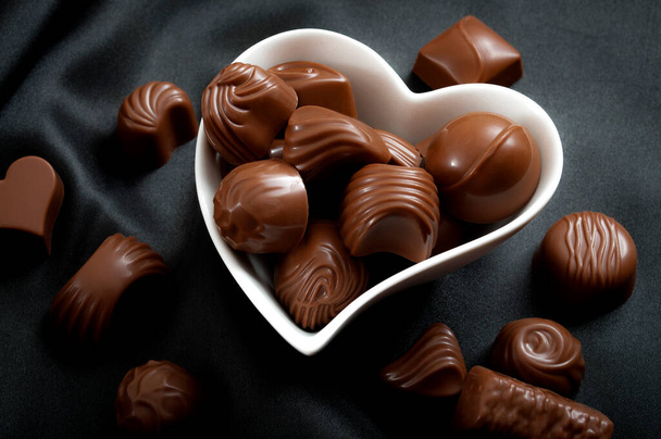 Regalos dulces románticos para el concepto del Día de San Valentín con un primer plano en una caja en forma de corazón llena de chocolates praliné o trufas de chocolate en seda negra o satén rodeado de pralinés caídos
 - Foto, Imagen