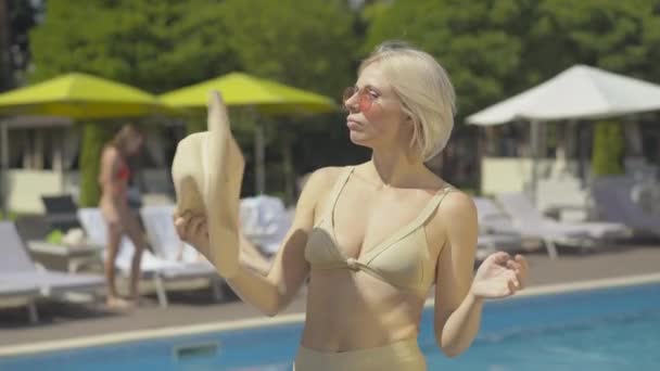 Middle shot of confident blond woman in golden suit waving sun hat at poolside. Portrait of beautiful adult Caucasian lady resting at luxurious summer resort. Relaxed slim female tourist on vacations. - Metraje, vídeo
