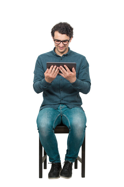 Jovem estudante do sexo masculino, sentado em uma cadeira, usando um tablet PC para entretenimento. Conceito de educação moderna, trabalhando e estudando em casa. Homem feliz vestindo óculos olhando para a tela do computador
. - Foto, Imagem