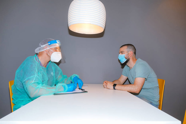 Travailleur médical avec écran facial assistant patient assis dans une clinique moderne. Discuter des symptômes et prendre des notes - Photo, image