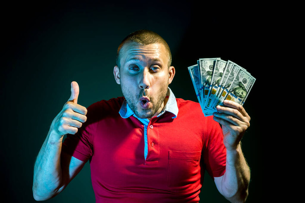 A happy Man with a fan of hundred dollar bills poses on a dark green background - Фото, изображение