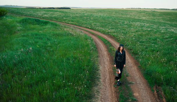 A young woman of 25-30 years old with curly red hair, in a black leather jacket and a tiara with feathers walks along a country road. - Foto, immagini