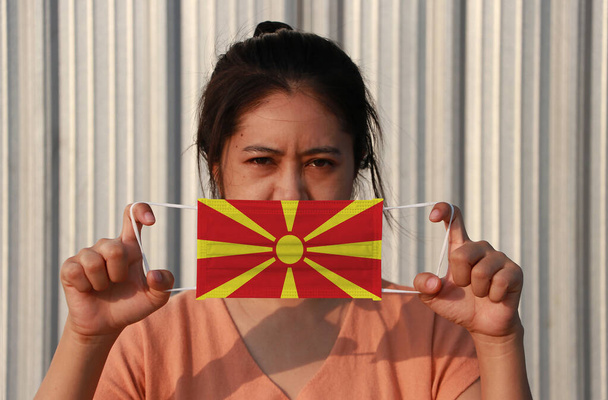 Kobieta z flagą Macedonii na masce higienicznej w ręku podniosła twarz na szarym tle. Drobne cząstki lub korona wirusa lub ochrona Covid 19. Koncepcja zwalczania chorób. - Zdjęcie, obraz