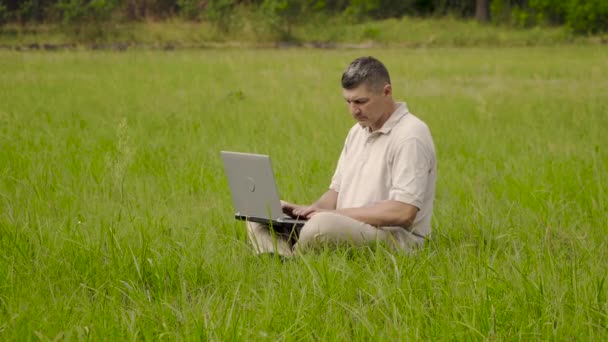 Serbest İşçi, çimenlikteki çimlerde otururken uzaktan kumanda için dizüstü bilgisayar kullanıyor. - Video, Çekim