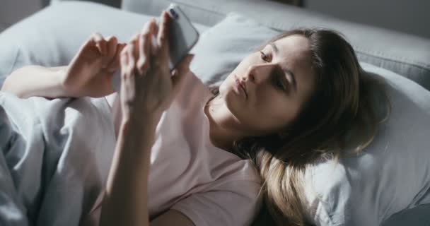 Top view on Caucasian young pretty woman lying in bed early in morning and texting message on smartphone. View from above on beautiful girl tapping and scrolling on mobile phone in bedroom. - Кадри, відео