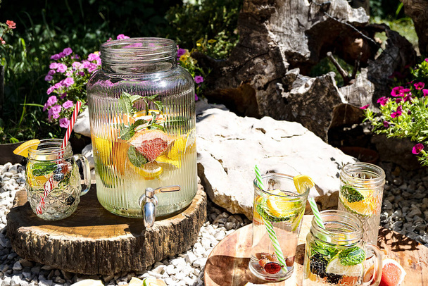 Homemade lemonade with lemon, lime, orange, grapefruit and mint in a large glass jug with a tap and cocktail mugs stands on a wooden stand in the open air in the garden. The concept of drinks, relaxation. - 写真・画像