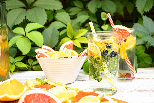 Mineral water with fruits and berries in a glass stands on a wooden table against the background of green foliage in the open air. Near a white bowl with juicy slices of fruit (orange, lime, grapefruit). - 写真・画像