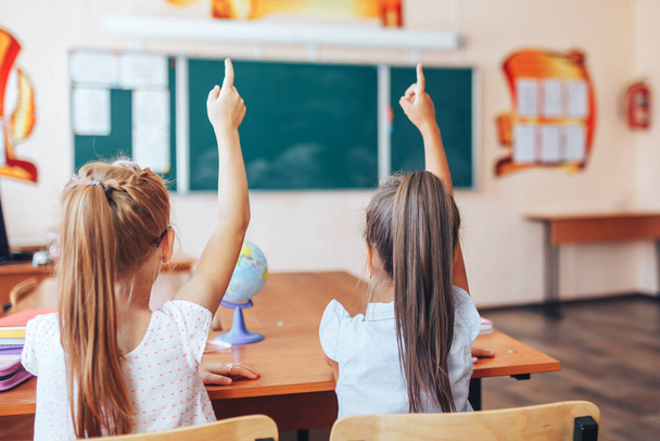 Δύο μαθητριούλες κάθονται σε ένα γραφείο σε μια σχολική τάξη και σηκώνουν τα χέρια τους. - Φωτογραφία, εικόνα
