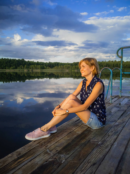 Een jong blank meisje met blond haar in spijkerbroek en een shirt zit op het meer. Zomer avond. Het concept van een ontspannen vakantie, de schoonheid van de natuur. - Foto, afbeelding