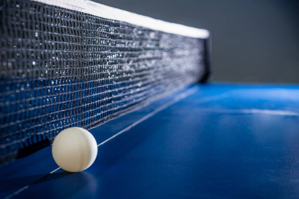 Close-up een witte bal op de blauwe ping pong tafel met een zwart net, Tafeltennis paddle is een sport competitie apparatuur indoor activiteit en oefening voor concept achtergrond - Foto, afbeelding