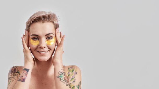 Beauty portrait of half naked tattooed woman with short hair smiling at camera, caring for her face, wearing under eye patches for dark circles isolated over light background - Photo, Image