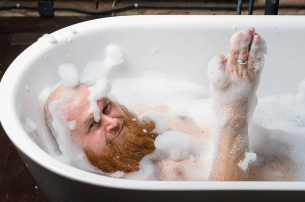 泡風呂に入る面白い赤い髭のはげ男のクローズアップ肖像画。スパの治療で陽気ないたずらな男。華やかな女の子のパロディー. - 写真・画像