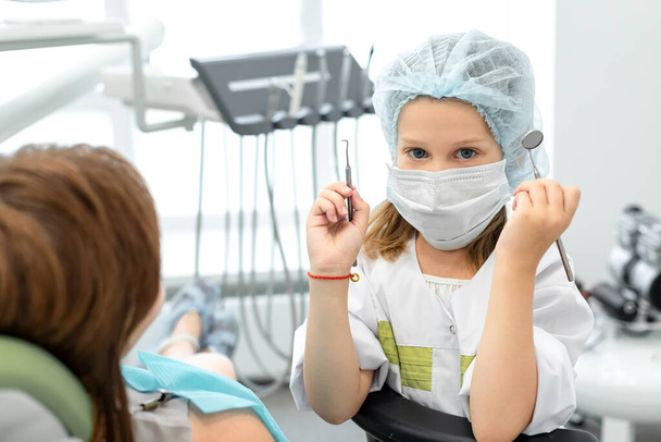 Καυκάσια επτάχρονη που έπαιζε στον οδοντίατρο. Παιδί κρατώντας οδοντιατρικά όργανα στα χέρια, γυναίκα οδοντίατρος απεικονίζει έναν ασθενή - Φωτογραφία, εικόνα