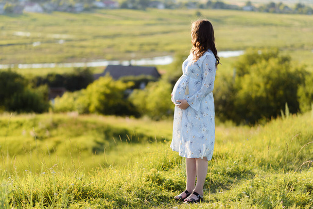 Close up of an expecting pregnant woman wearing a blue dress and holding her belly and relaxing outdoor - Fotó, kép