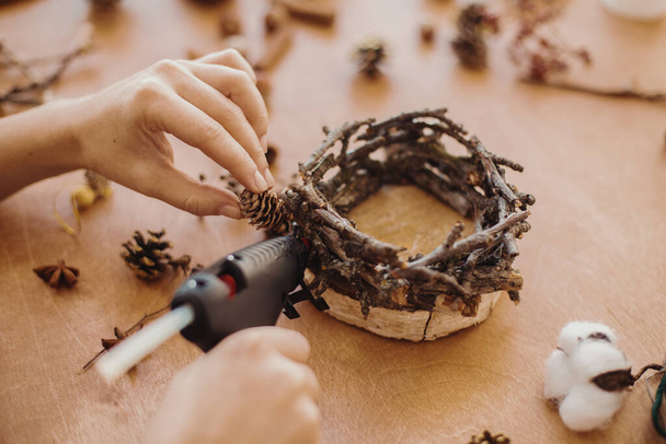 Hands holding glue gun and making rustic festive decoration from  pine cones, branches, and cotton on wooden table. Holiday workshop. Making rustic christmas candle holder. - Фото, зображення