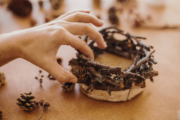 Florist making rustic christmas candle holder. Hands holding pine cones and branches and making rustic festive decoration on wooden table. Holiday workshop - Foto, Imagem