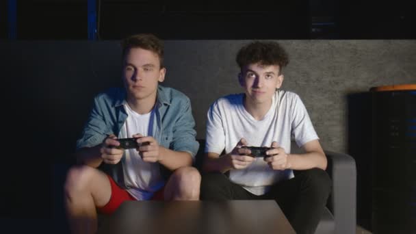 Dwóch młodych facetów siedzi na kanapie przed telewizorem i jest podekscytowanych grą w konsolę do gier. Jeden przegrywa, drugi wygrywa. - Materiał filmowy, wideo