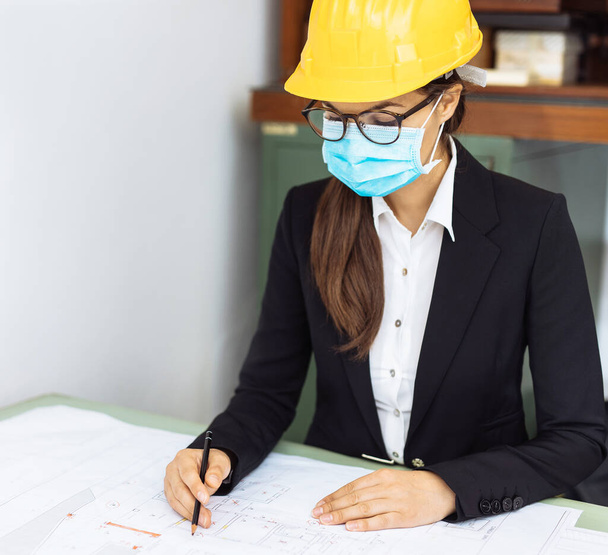 The young female architect draws a sketch with a pencil. Young designer ledy planning at the office desk. Respect all safety measures wearing a helmet and mask - Photo, Image