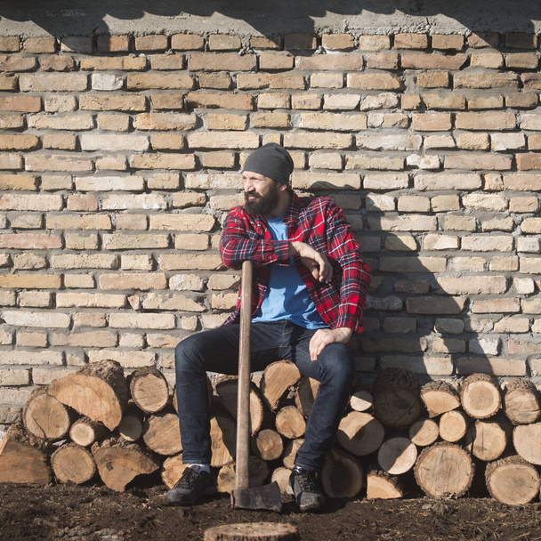 Уставший лесоруб сидит и отдыхает перед кирпичной стеной, рабочий в сельской местности. - Фото, изображение