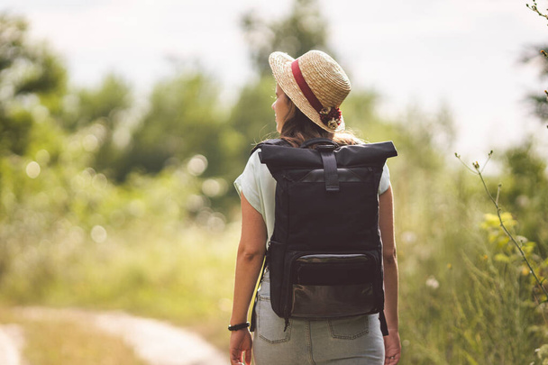 Stylish fashionable hipster traveler woman in hat with brim and roll top type backpack walking in nature. Travel and wanderlust concept. Exploring nature, hot summer day. Outdoor activity. Wanderlust. - Photo, Image