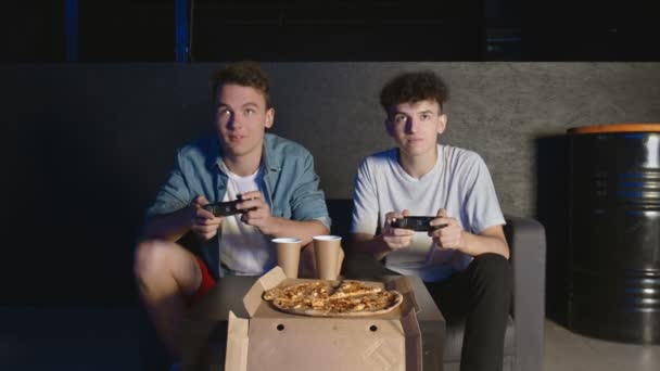 リビングルームで二人の友人はソファに座ってビデオゲームをプレイし、ピザを食べる. - 映像、動画